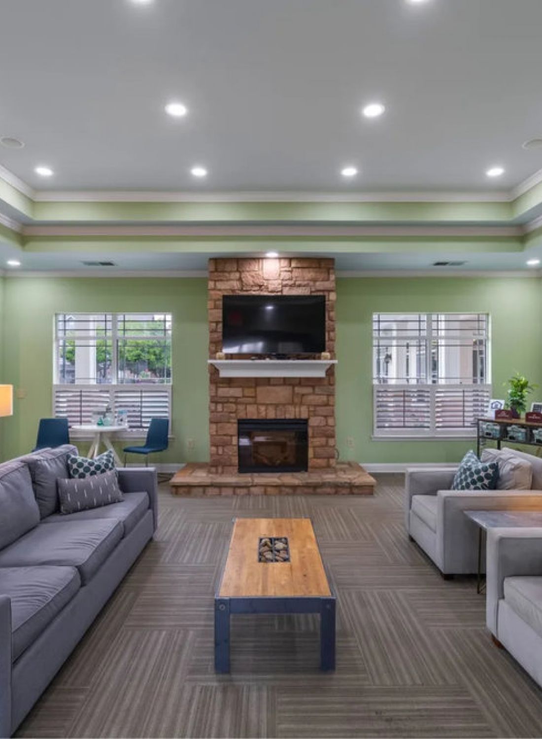 Modern lounge with gray sofas, a stone fireplace, TV, coffee tables, lamps, and green walls with large windows.