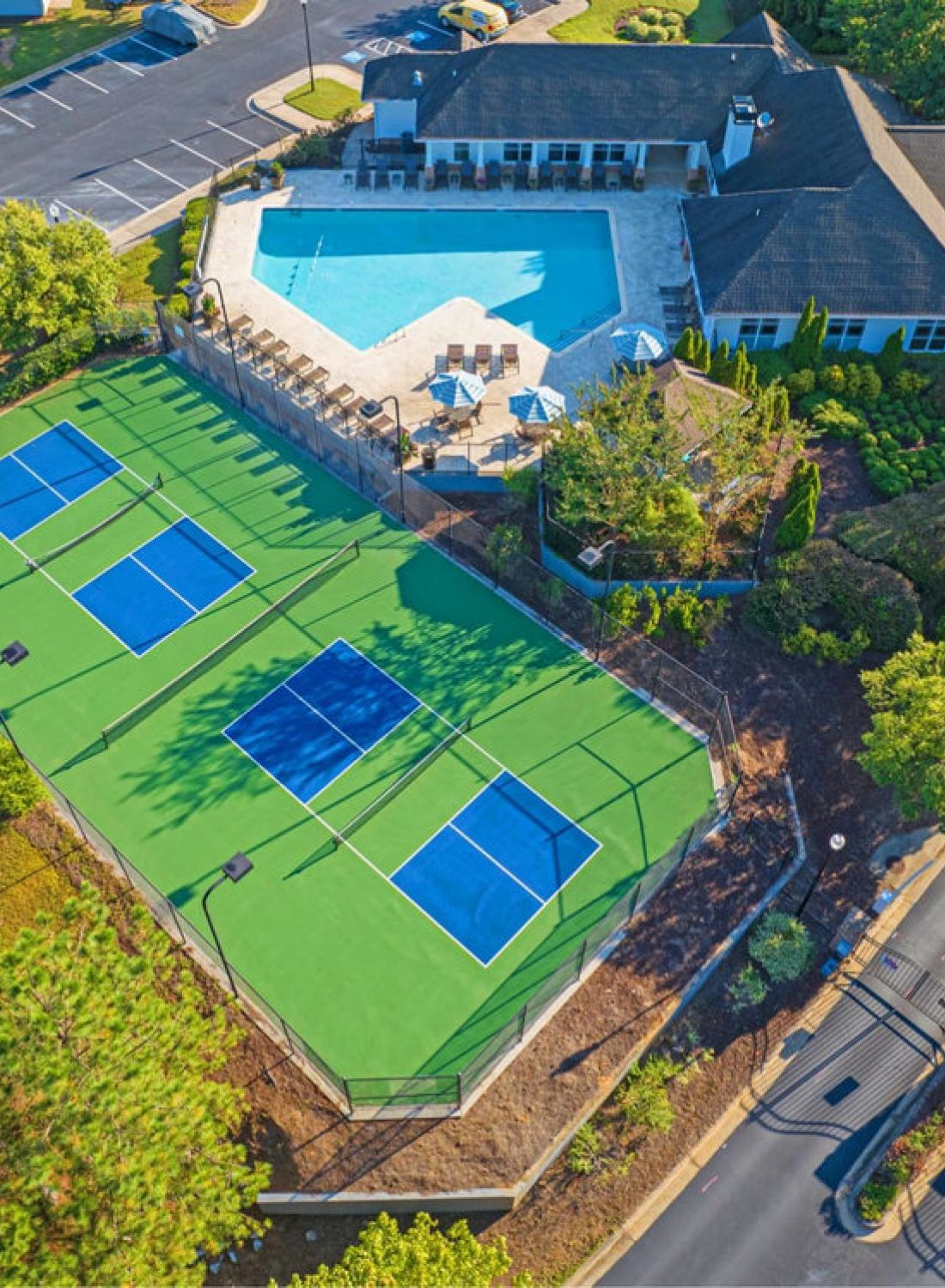 Aerial view of tennis courts, swimming pool, and clubhouse surrounded by trees and parking lot.