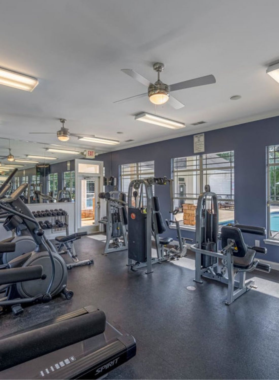 Modern fitness room with cardio and weight machines, large mirrors, and windows overlooking a pool area.
