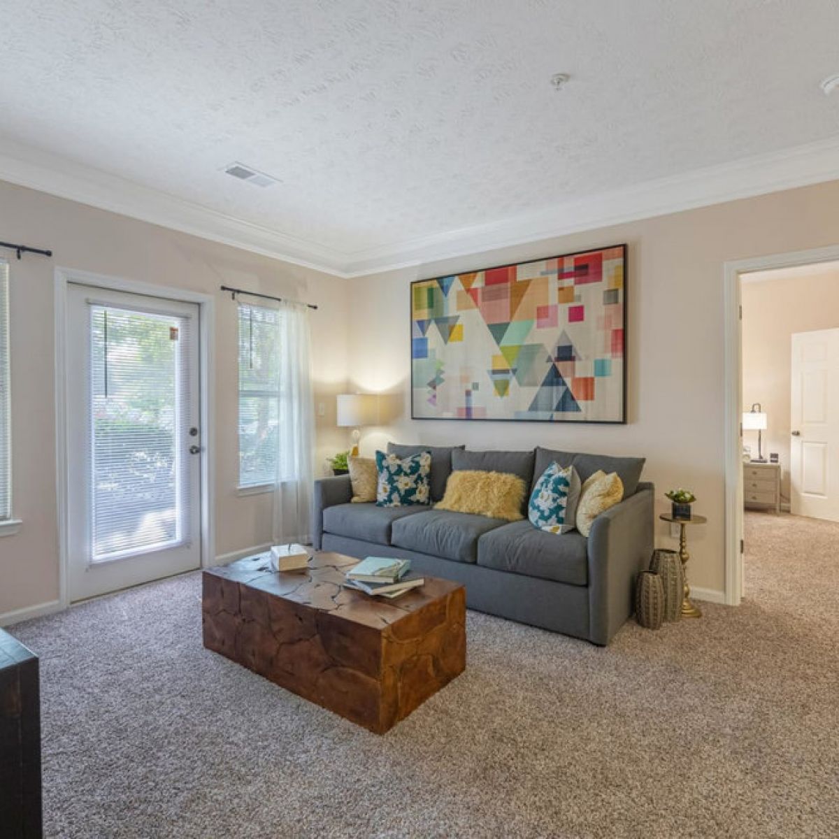 Bright living room with a gray sofa, abstract art, wooden coffee table, and open doorway to a bedroom.