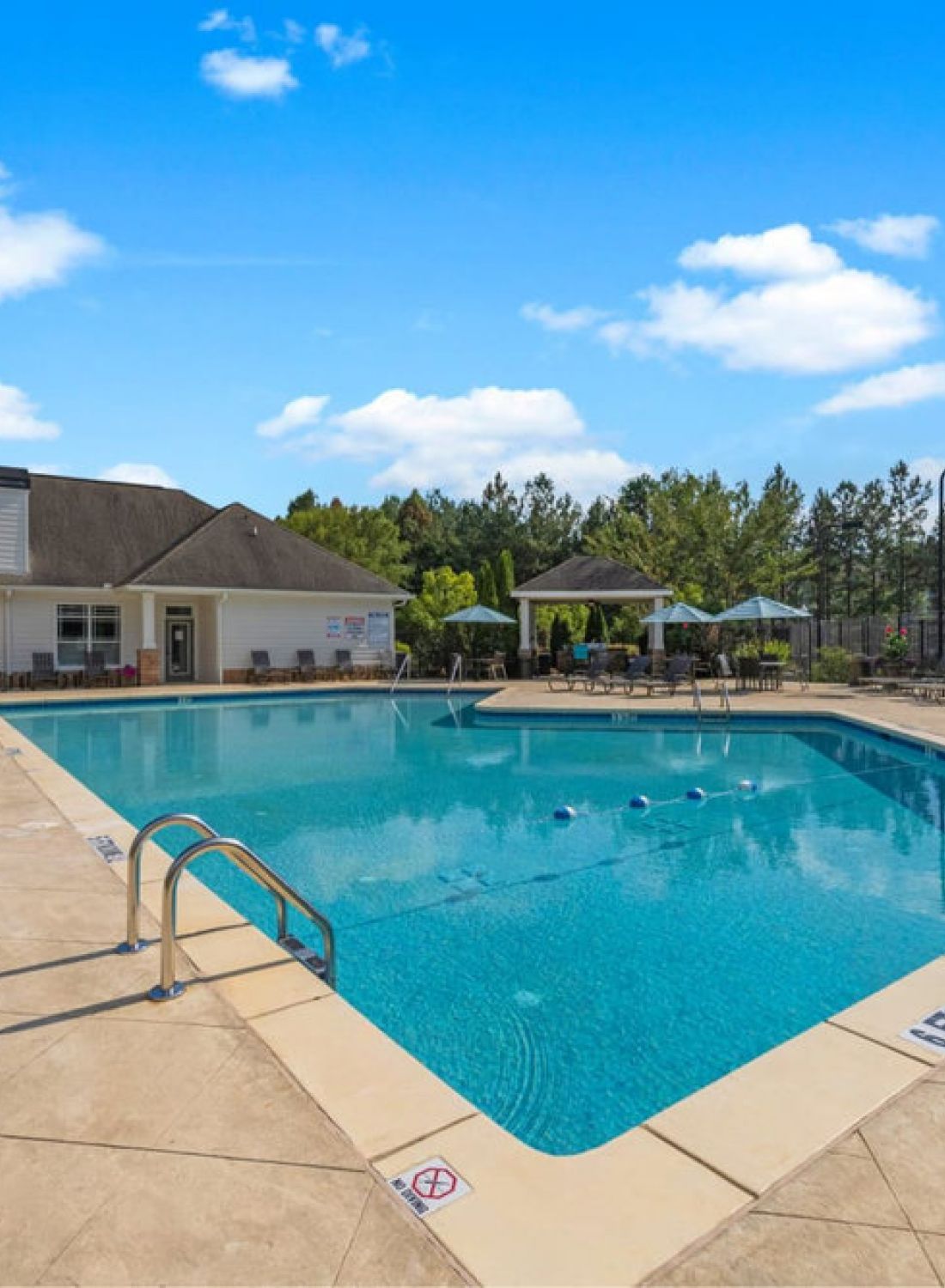 Outdoor swimming pool with lounge chairs and umbrellas, surrounded by trees under a blue sky with clouds.