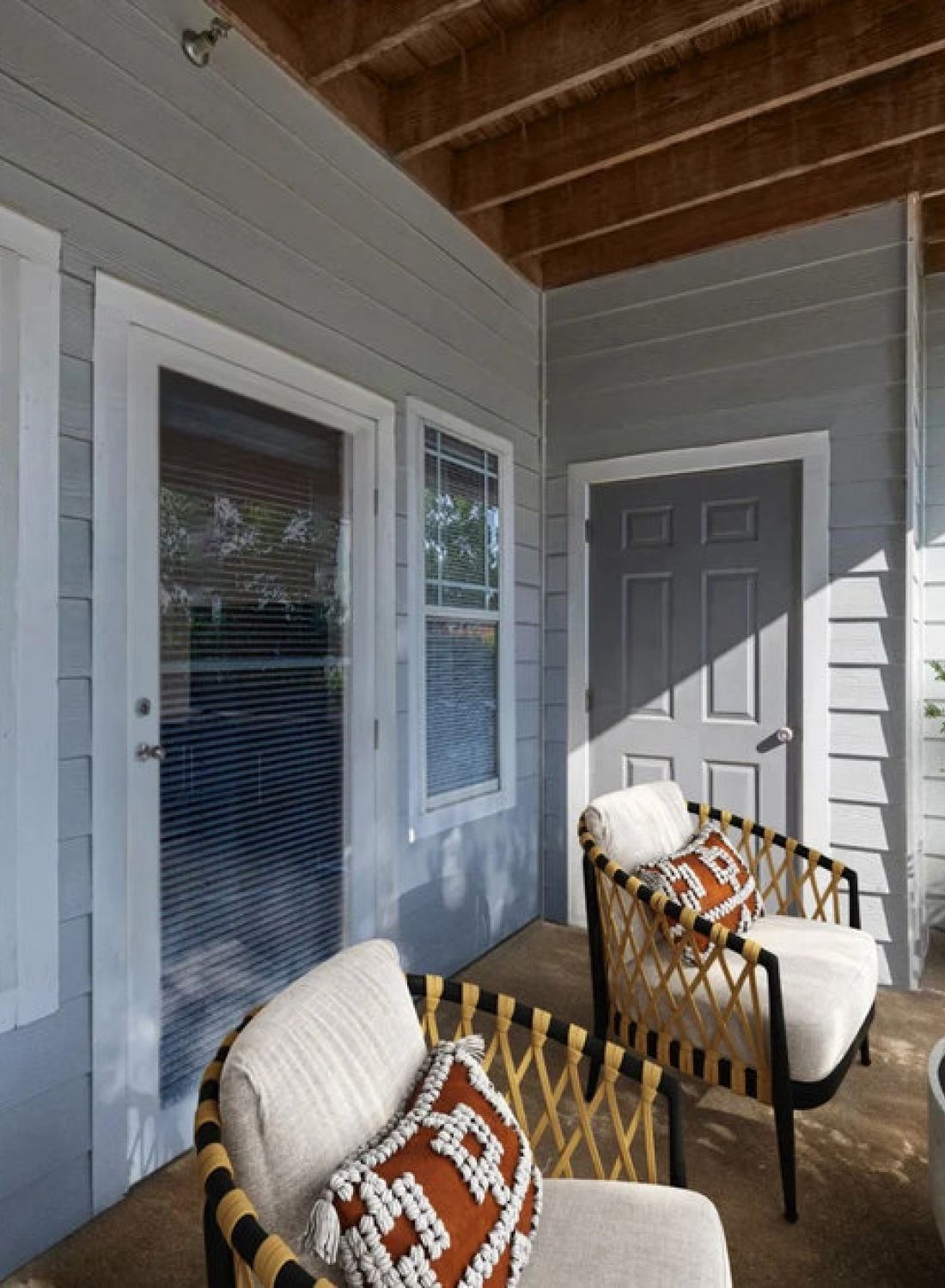 Covered patio with two cushioned chairs, a round table, potted plants, and gray siding on the building.
